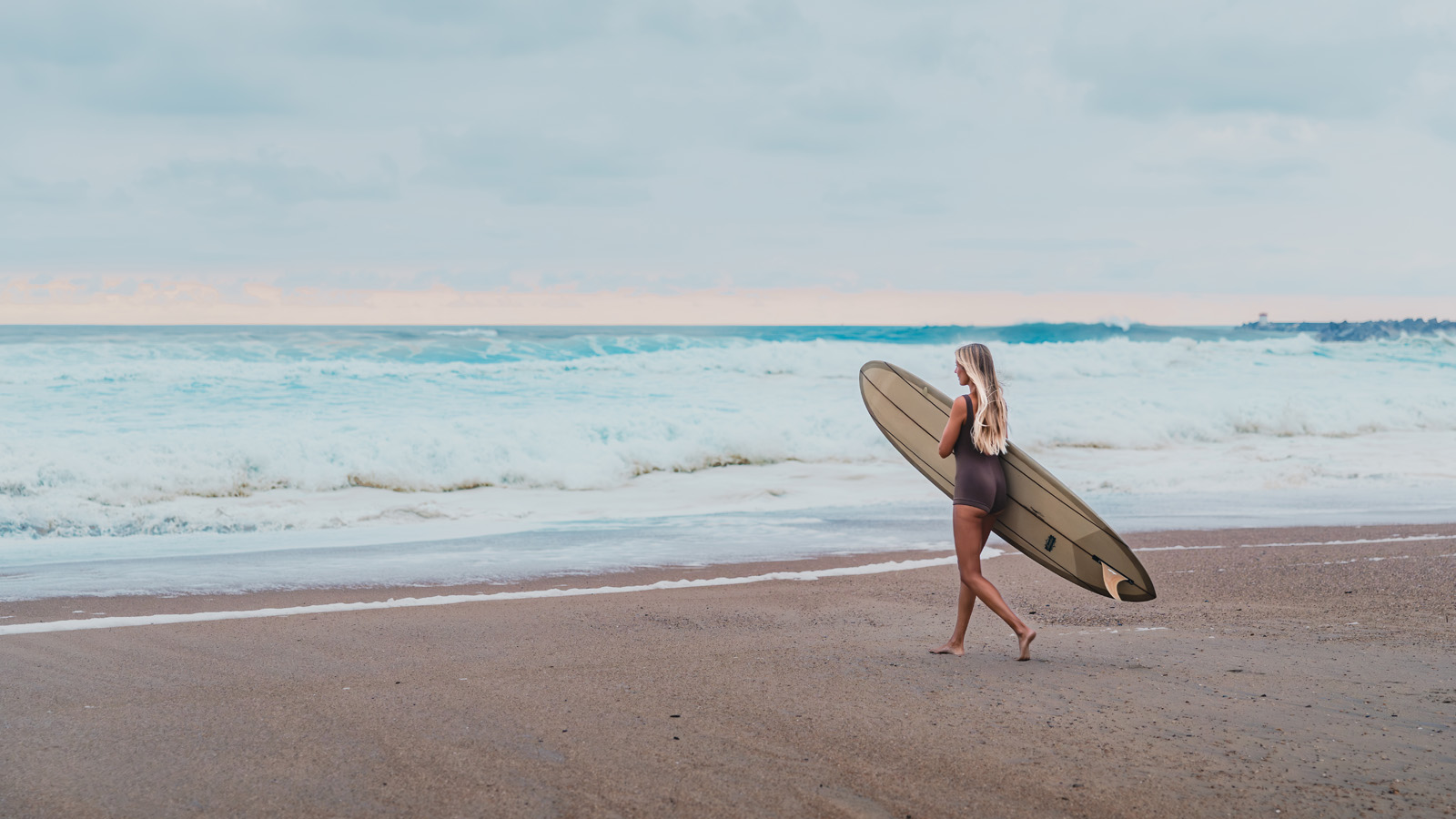 Blonde female surfer walking on a sandy beach carrying a longboard surfboard toward the ocean waves.