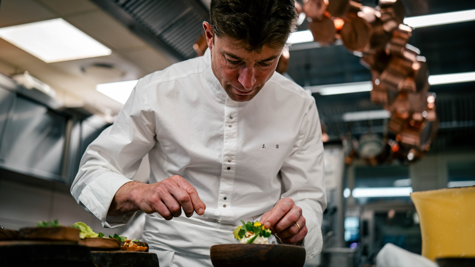 Michelin-starred chef Jean Sulpice, in a white chef's jacket with his initials 'J.S.', meticulously plating gourmet appetizers in a professional kitchen featuring hanging copper pots and a large wall clock.