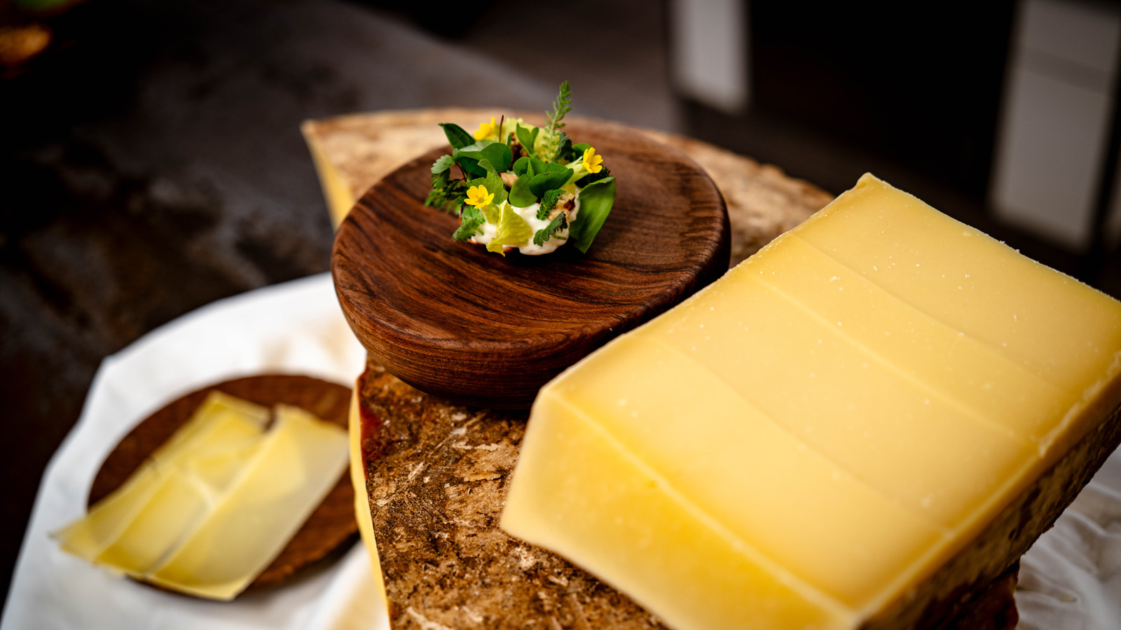 Close-up of a large block of firm yellow Alpine cheese alongside a gourmet appetizer garnished with herbs and yellow flowers on a small wooden plate.