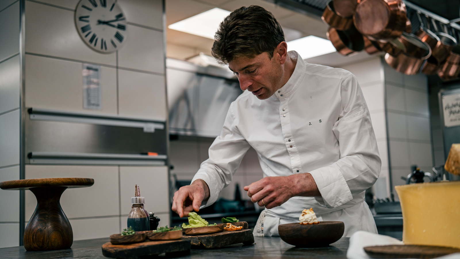 Michelin-starred chef Jean Sulpice, wearing a white chef's coat with his initials 'J.S.', meticulously garnishing a gourmet dish in a professional kitchen with copper pots hanging in the background.