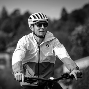 A man in a white and olive jacket and helmet riding a matte grey e-bike with a wooden crate of fresh herbs on the rear rack, passing in front of a rustic stone building with an arched wooden door.