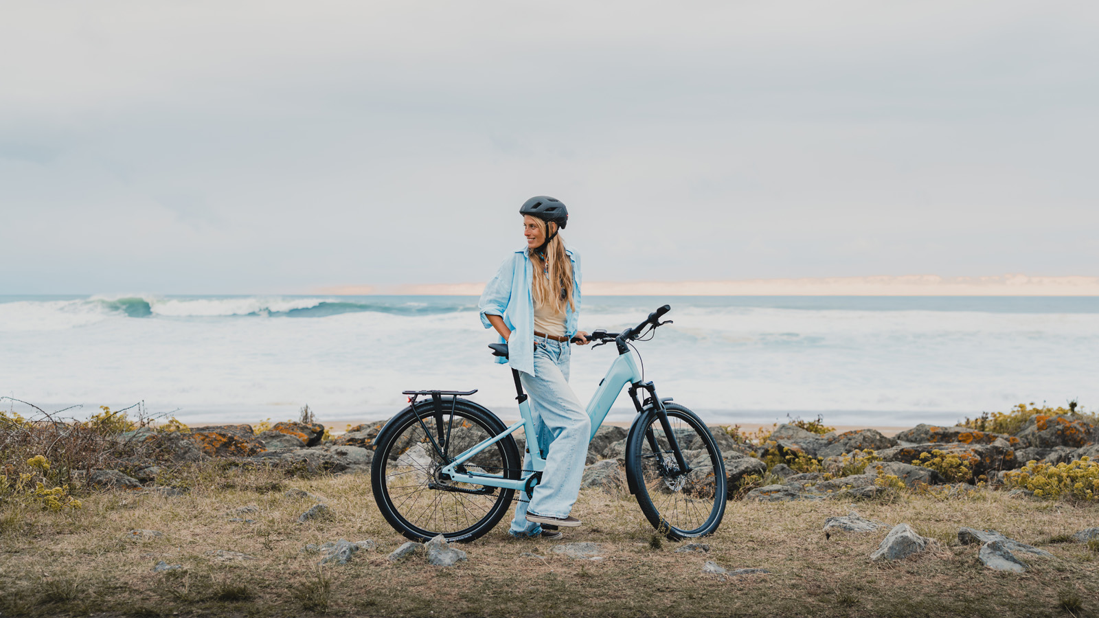 A woman in a blue shirt, light-wash jeans, and a dark helmet stands with a light blue step-through electric bike on a grassy, rocky bluff overlooking a vast, white-capped ocean under a cloudy sky.