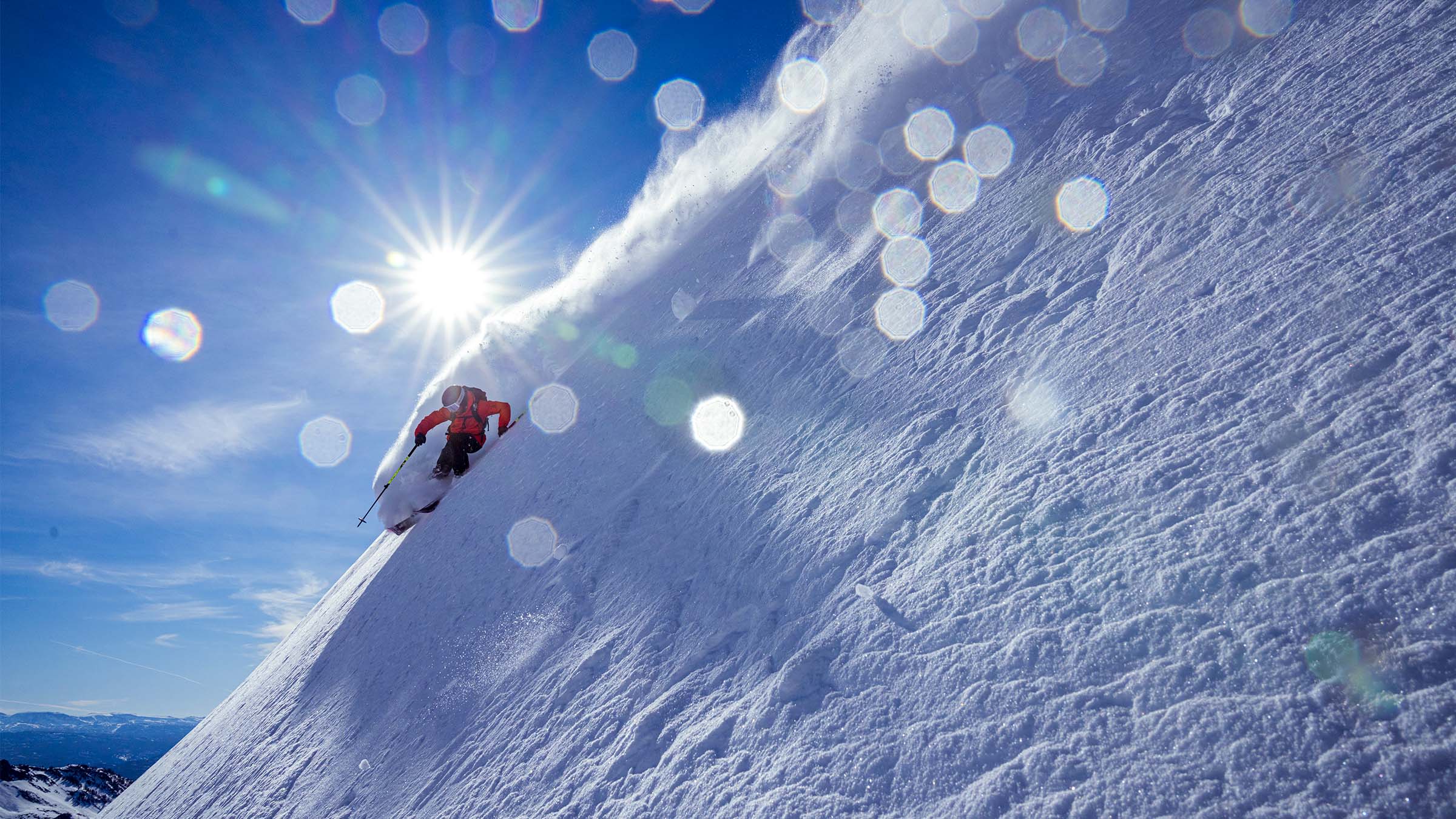 Sam Cohen skiing in a plume of pow while backlit with a sun burst and snow