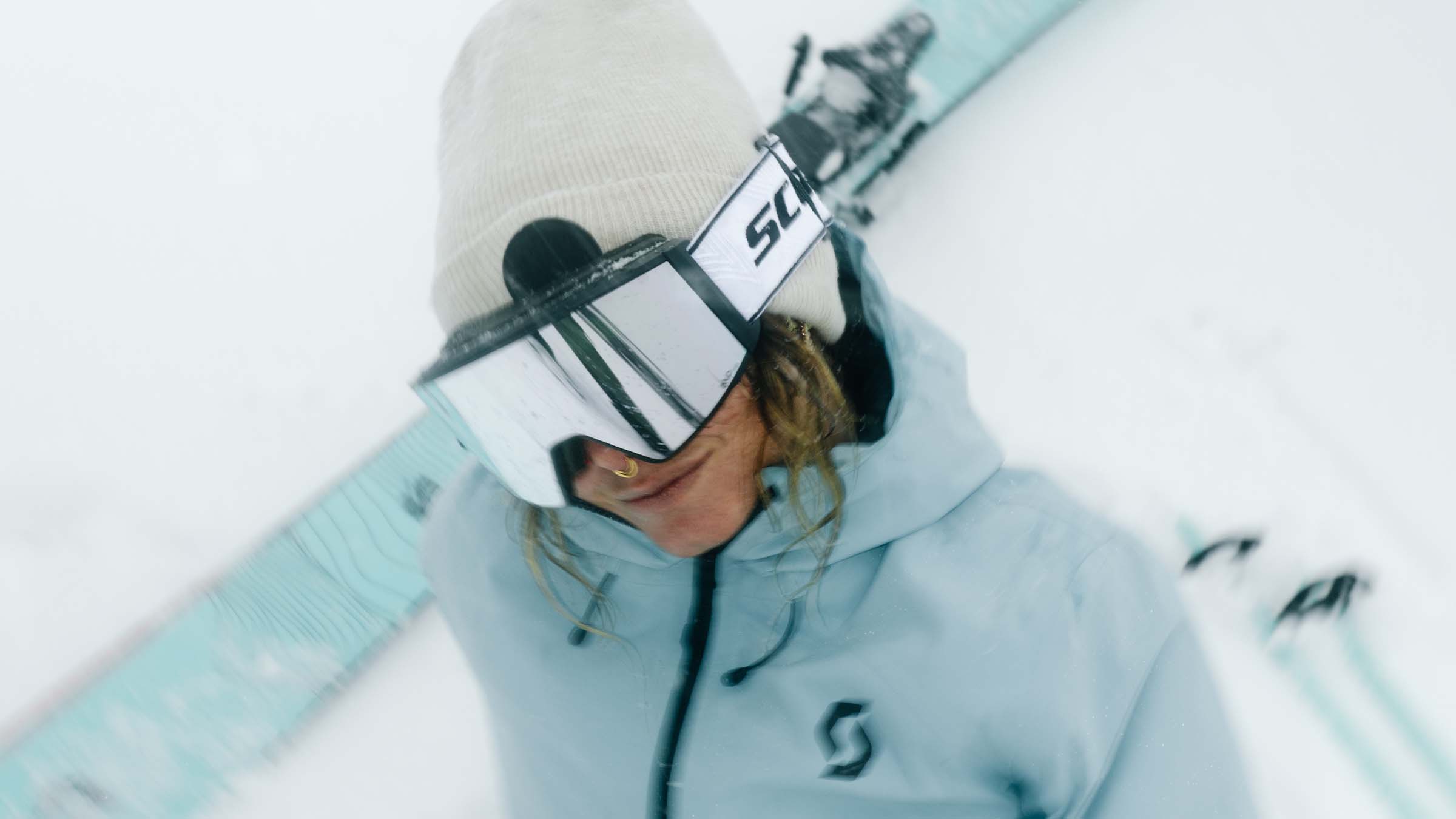 A women wearing SCOTT goggles with a white beanie while sitting in the snow.
