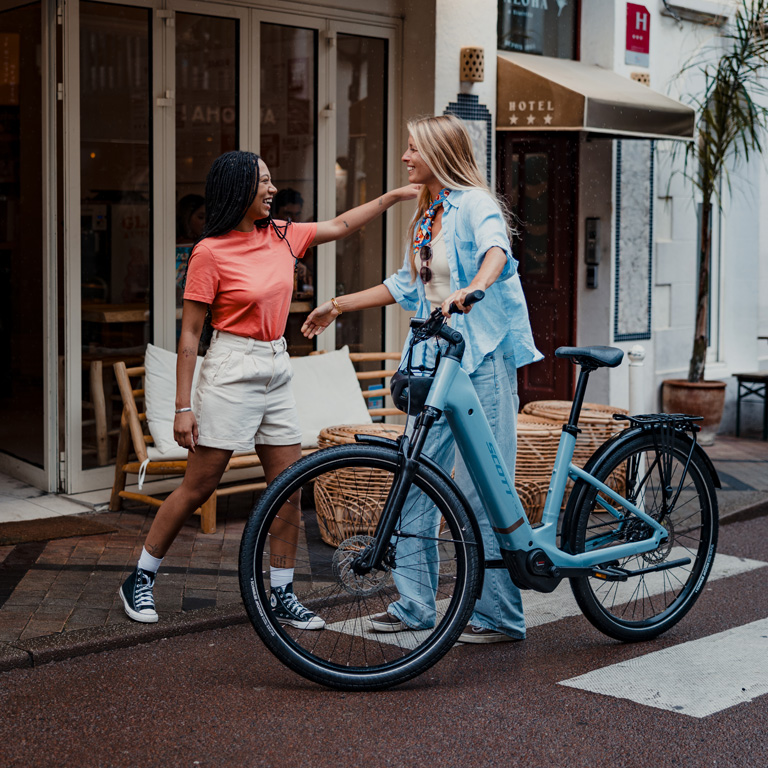 A woman with a light blue Scott electric bike greeting a friend outside a hotel cafe on a city street.
