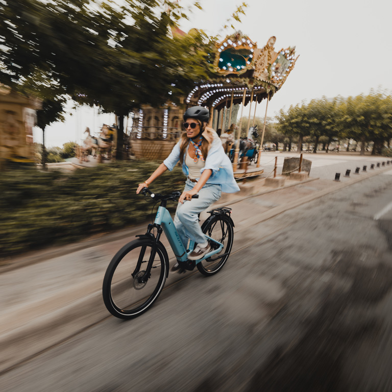 Woman riding a light blue Velotric Discover 2 step-through electric bike past a classic carousel in a park.