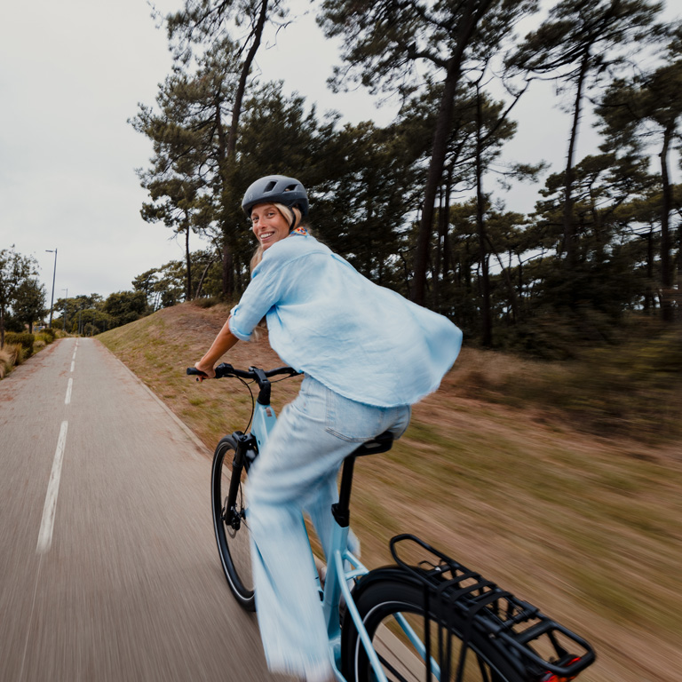 Smiling woman in light blue casual wear riding a light blue step-through electric bike on a paved forest trail.