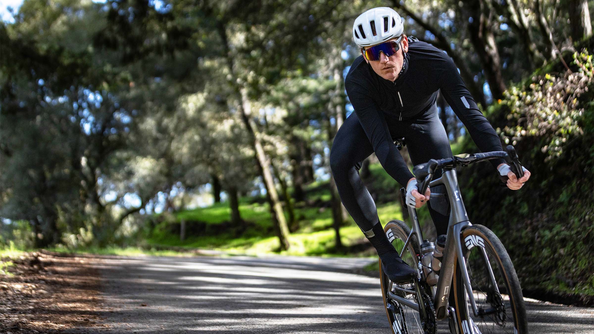 Brennan Wertz cornering at speed on a SCOTT Addict 10 road bike, wearing a white helmet and black kit, riding a shaded, tree-lined road.
