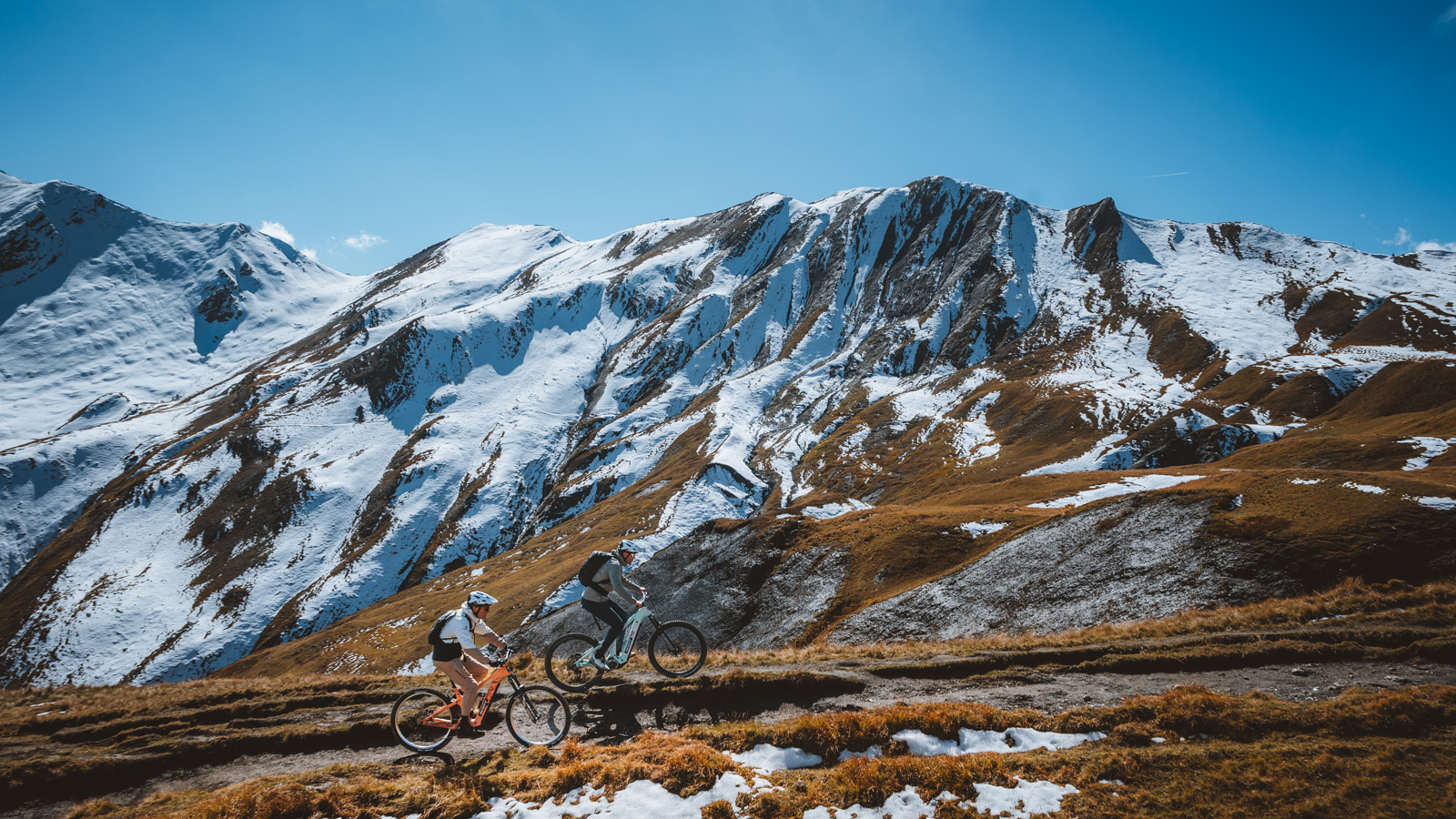 Two mountain bikers riding downhill on a narrow trail across an alpine meadow with autumn colors, dramatic clouds and rugged mountains in the background.