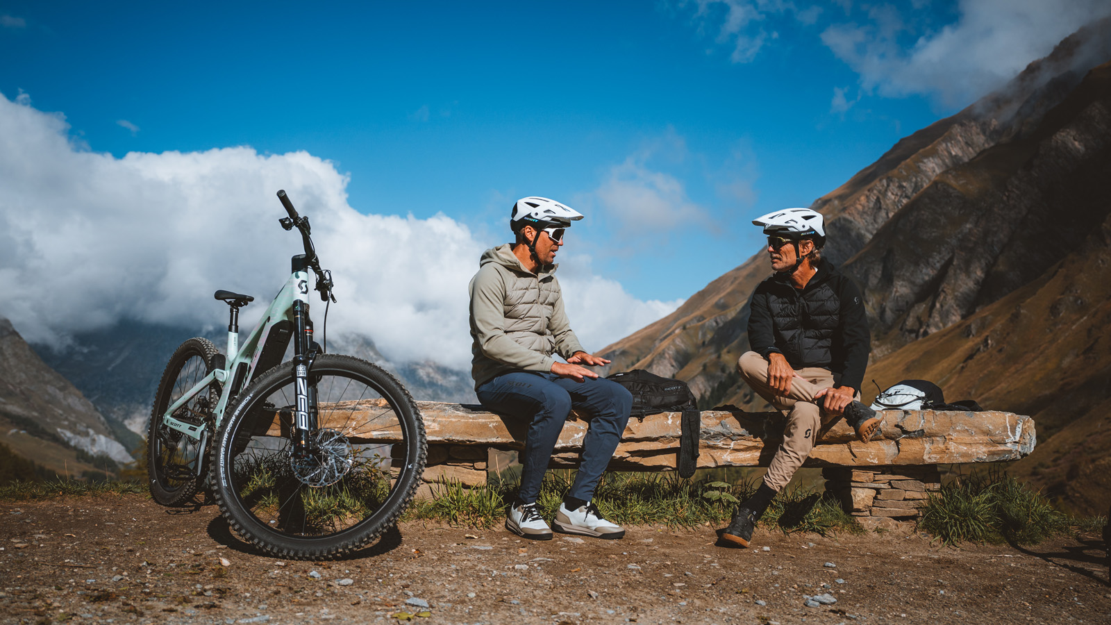 Two mountain bikers riding along a scenic alpine trail with snow-covered mountains and a clear blue sky in the background.