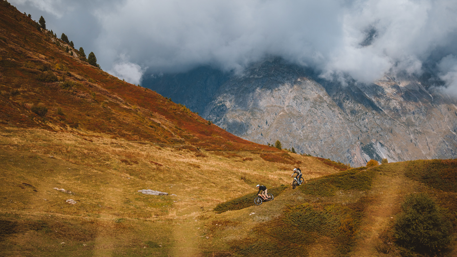 Two mountain bikers sitting on a stone bench talking during a break, with an e-mountain bike beside them and alpine peaks behind.