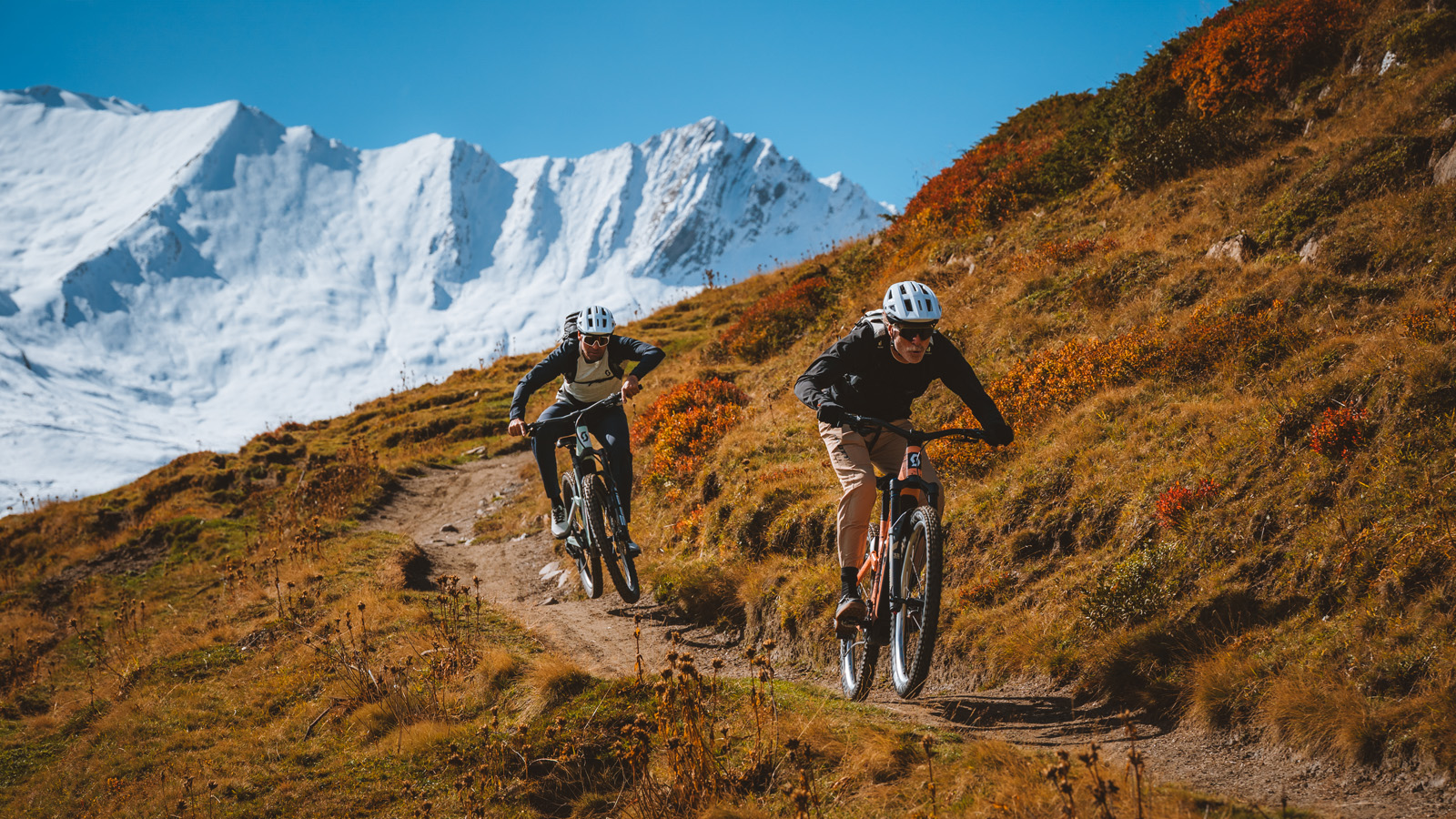 Two mountain bikers in helmets ride down a rugged trail on a grassy hillside, with snow-capped mountains in the background under a clear blue sky.