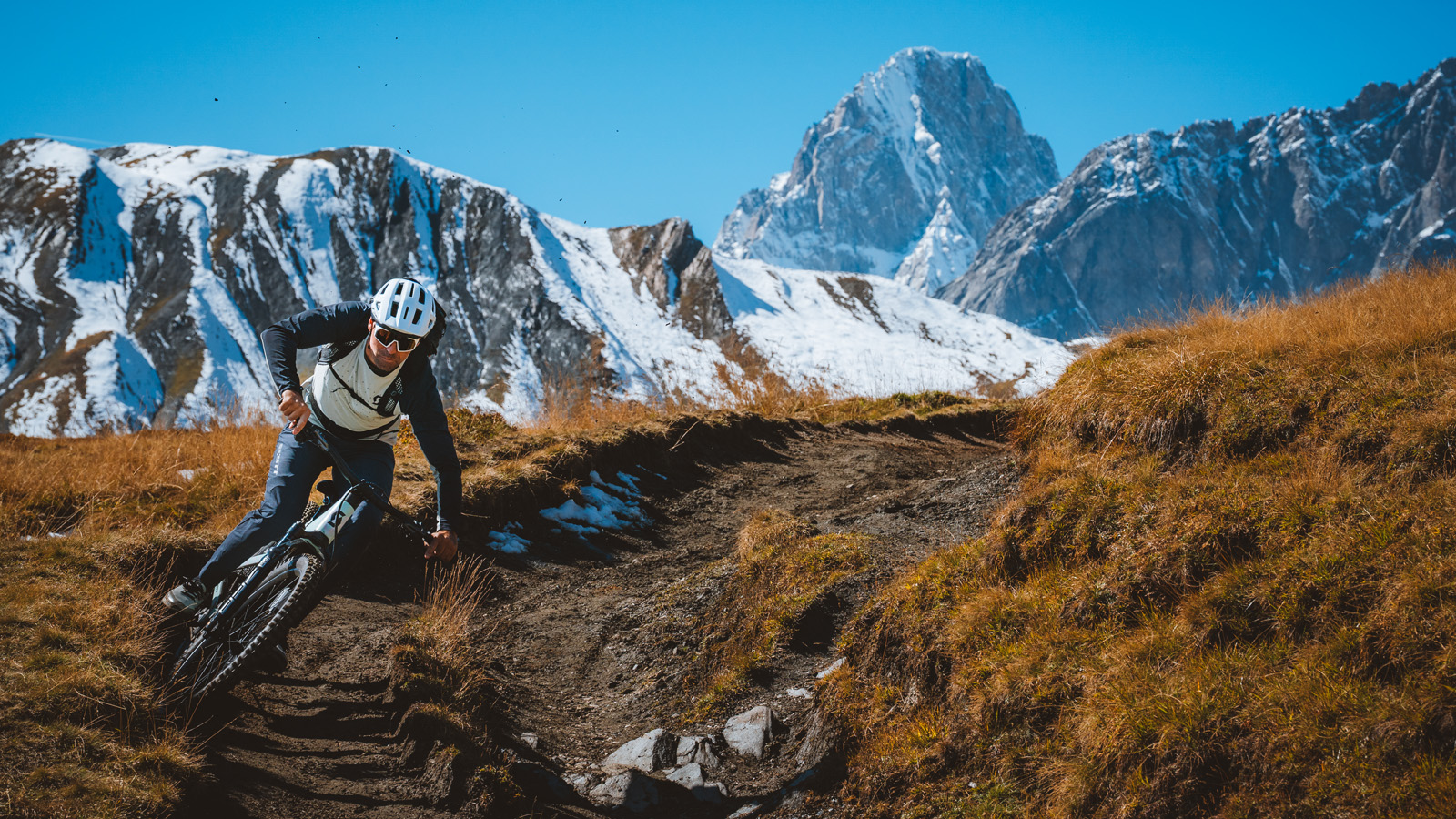 A cyclist in a white helmet rides down a dirt path on a mountain. Snow-capped peaks loom in the background on a sunny day, creating an adventurous vibe.