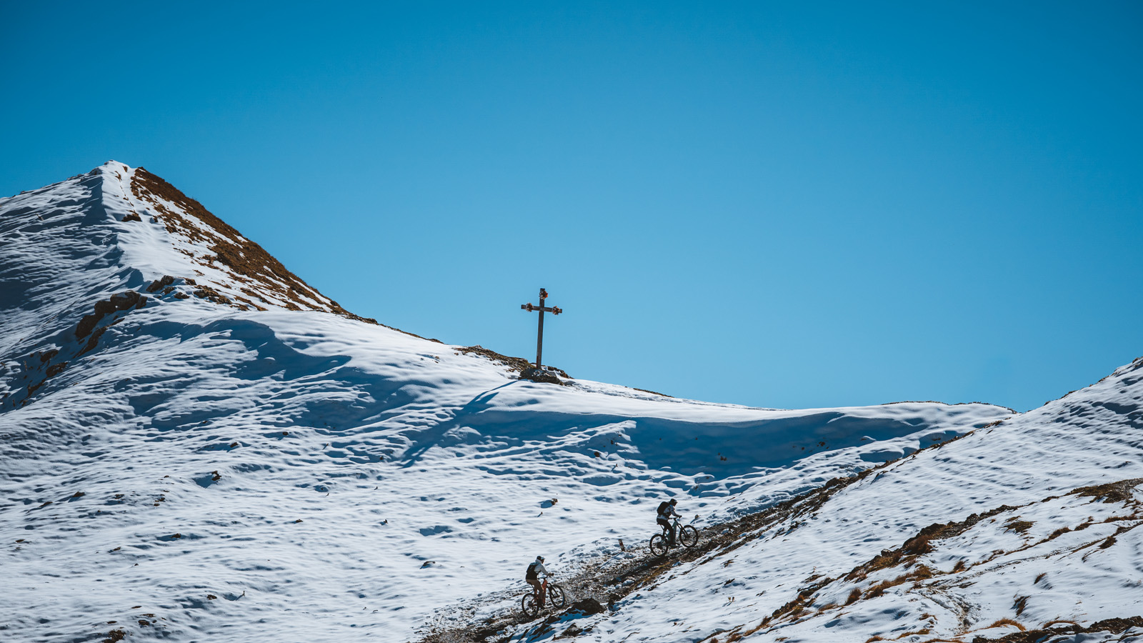 Two cyclists navigate a snowy mountain path under a clear blue sky. A cross stands prominently at the summit, evoking a sense of adventure and tranquility.