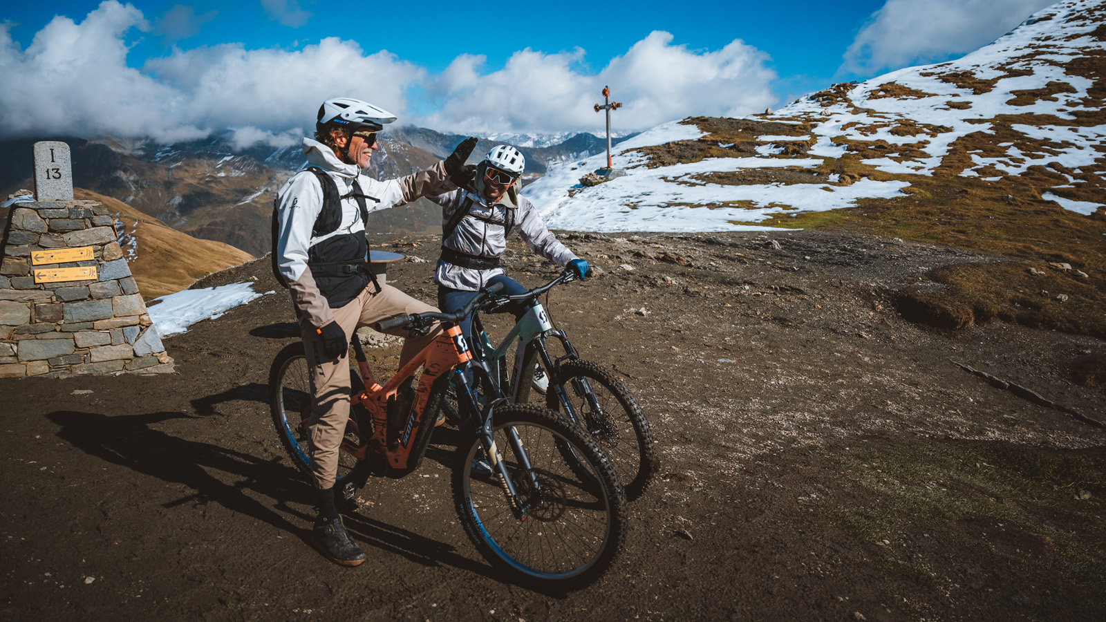 Two cyclists in winter gear and helmets stand with mountain bikes on a snow-dusted trail. They appear cheerful, set against a scenic mountain backdrop.