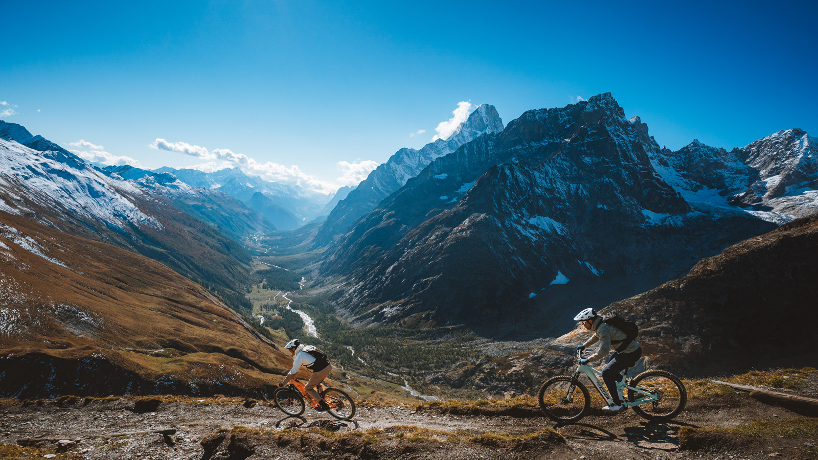Two mountain bikers ride along a rugged trail with a majestic mountain range in the background. The scene is bright and exhilarating, capturing the thrill of adventure.