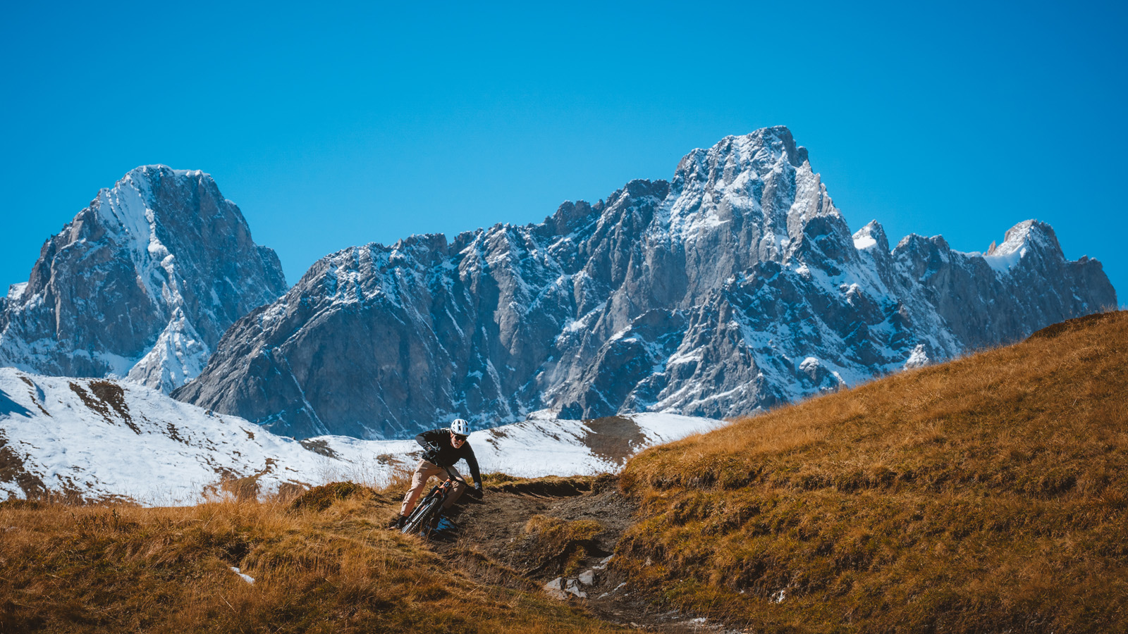 Mountain biker riding a singletrack trail through alpine grassland with snow-covered jagged mountain peaks in the background under a clear blue sky, extreme outdoor adventure landscape, high-altitude mountain biking in a dramatic alpine environment.