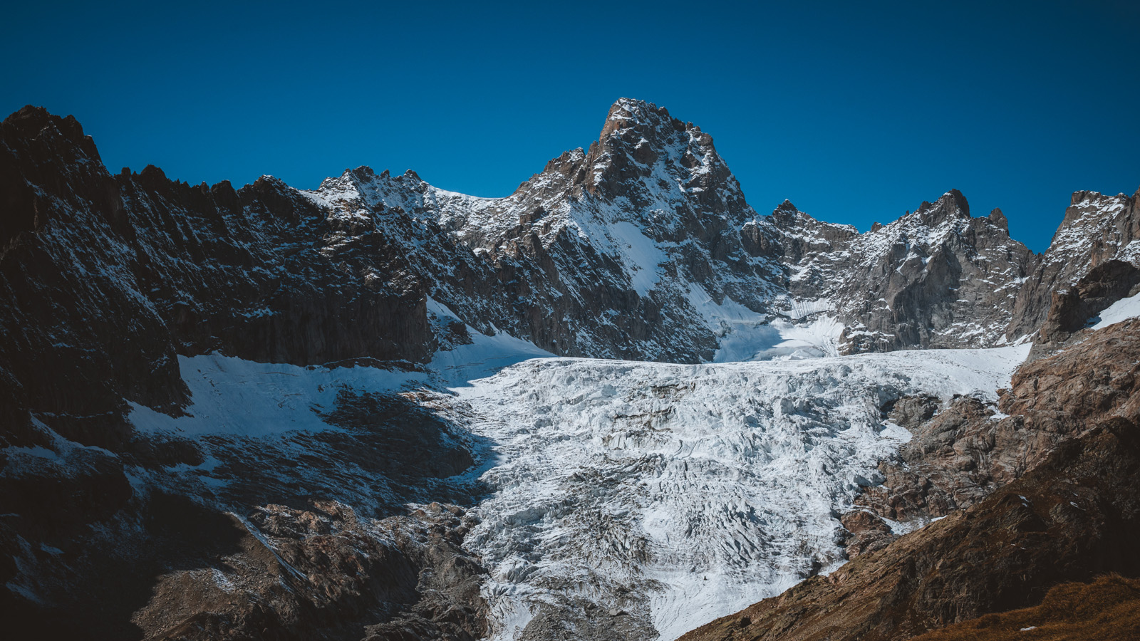 Massive glacier flowing between rugged rocky mountain peaks with patches of snow and ice under a deep blue sky, dramatic alpine glacier landscape, high-altitude wilderness scenery with glacial ice and steep cliffs.