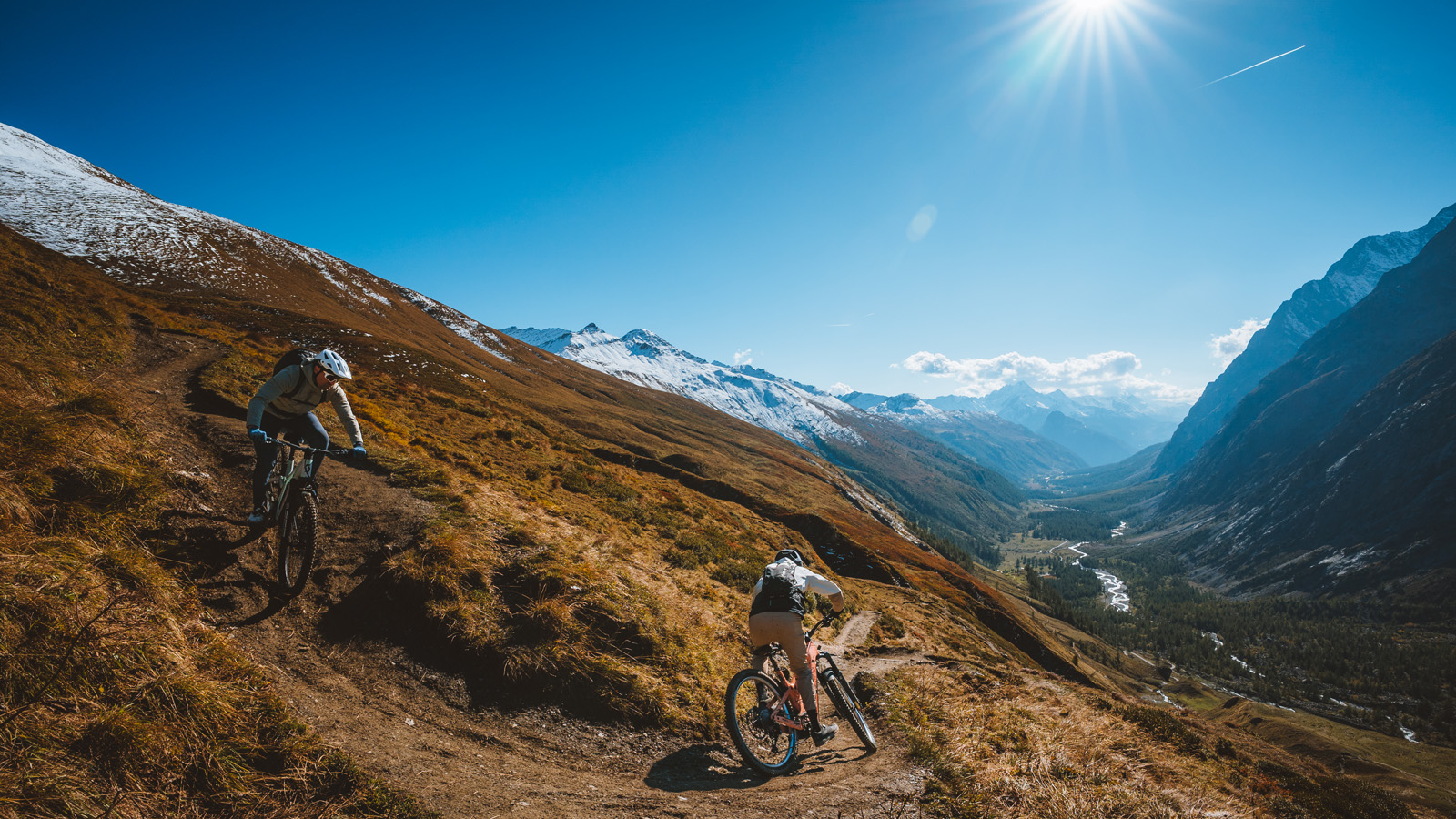 Two cyclists ride mountain bikes down a dirt trail on a sunlit, rugged hillside with snow-capped peaks and a valley in the background.