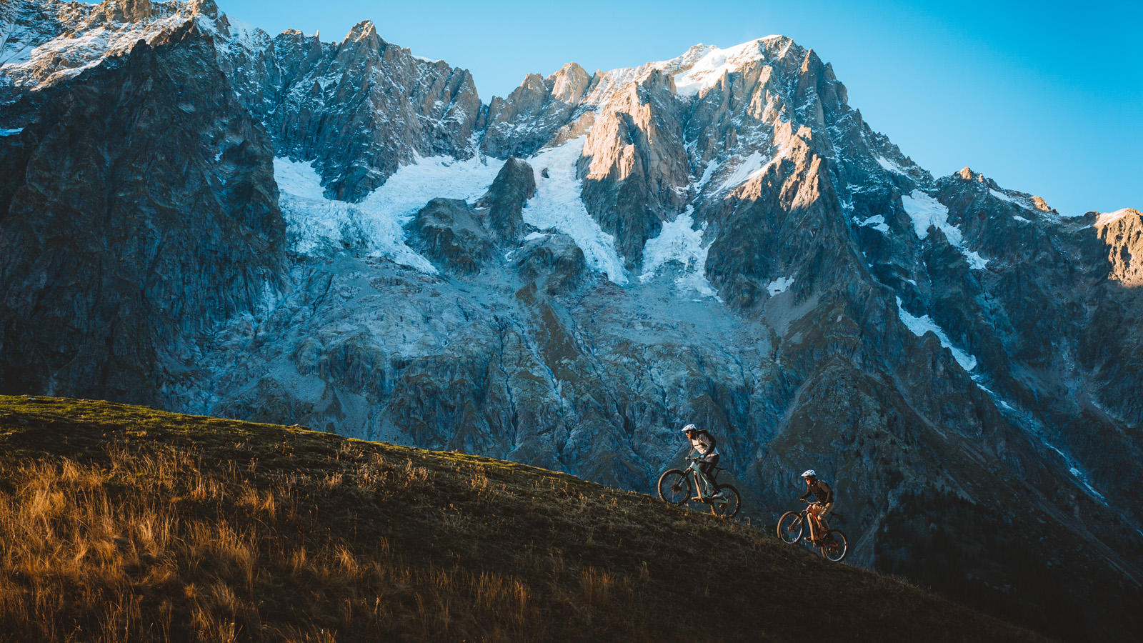 Two cyclists ride uphill on a grassy slope against a backdrop of majestic, sunlit snow-capped mountains under a clear blue sky.