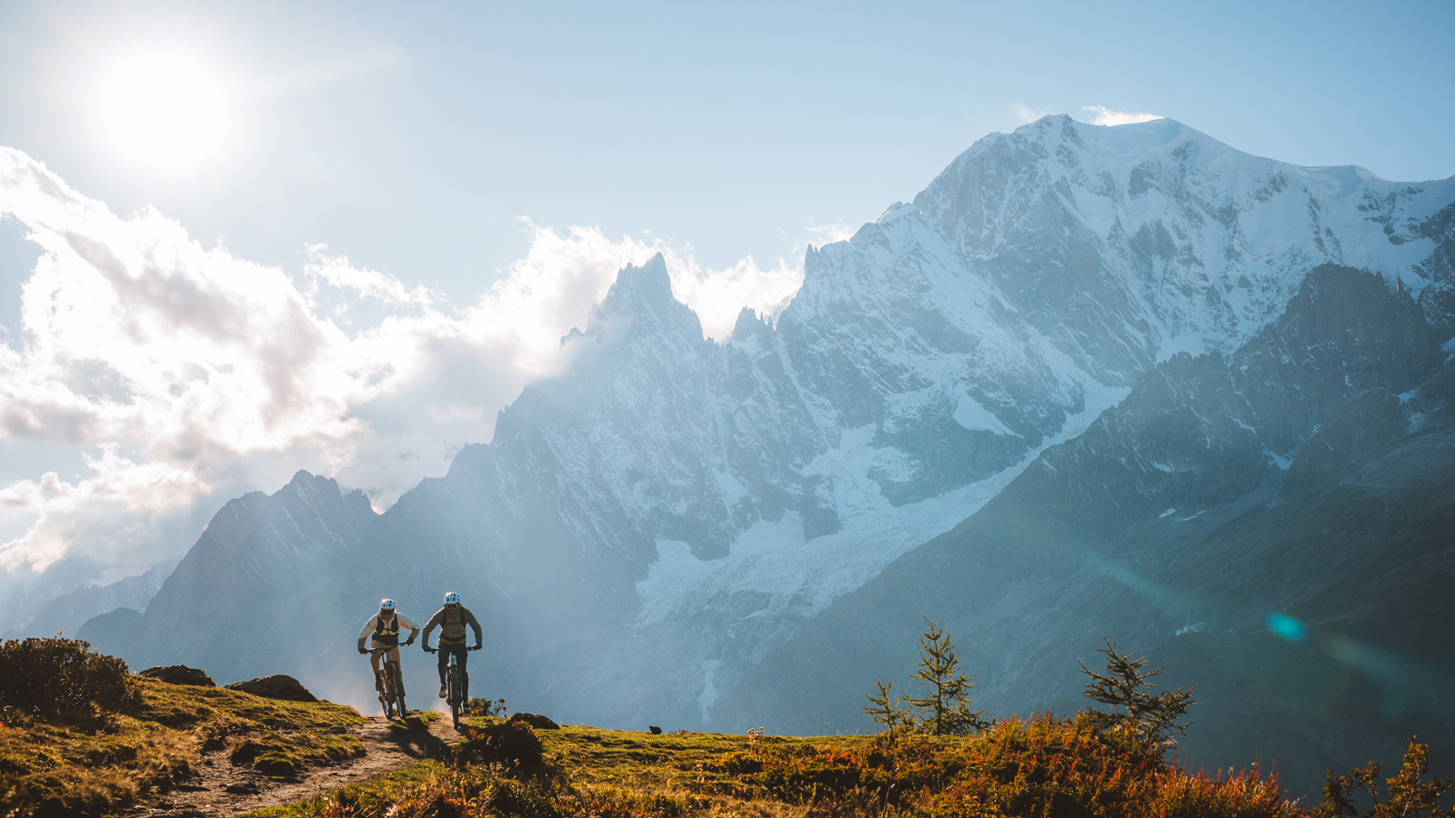 Two cyclists ride on a mountain trail under a bright sun, near towering, snow-covered peaks. The scene conveys adventure and tranquility.