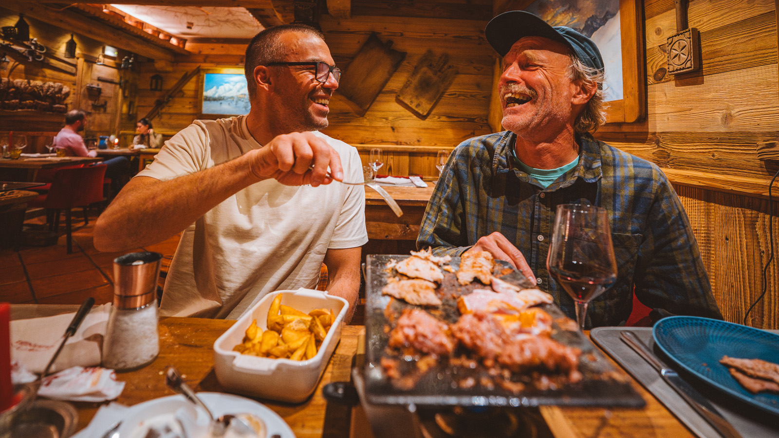 Two men share a joyful meal in a cozy wooden restaurant. They laugh heartily, sitting beside a sizzling platter of meat and a dish of fries, creating a warm, convivial atmosphere.
