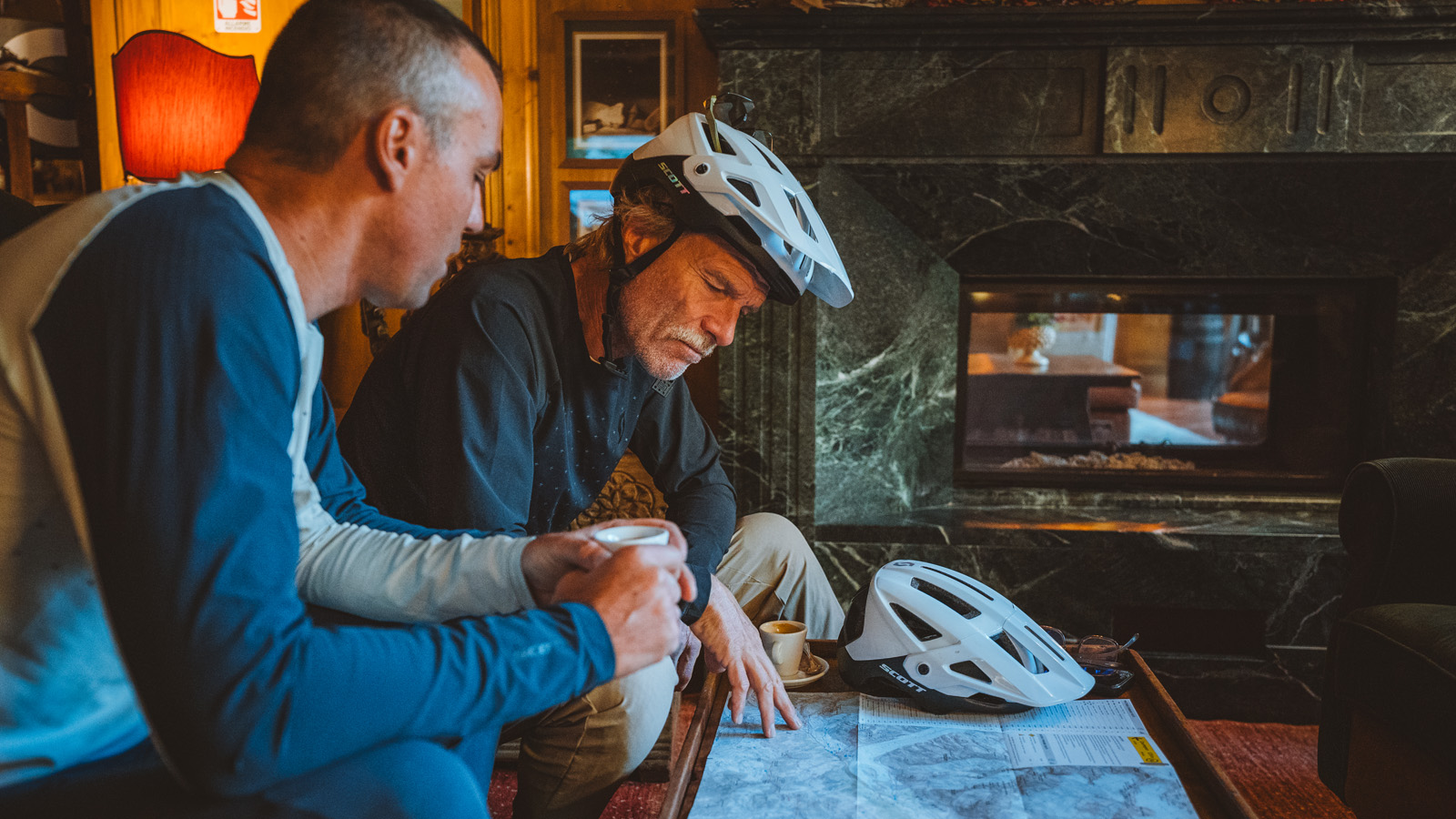 Two men with white helmets sit near a fireplace, studying a map on a table. One holds a coffee cup. The room has a cozy, focused ambiance.