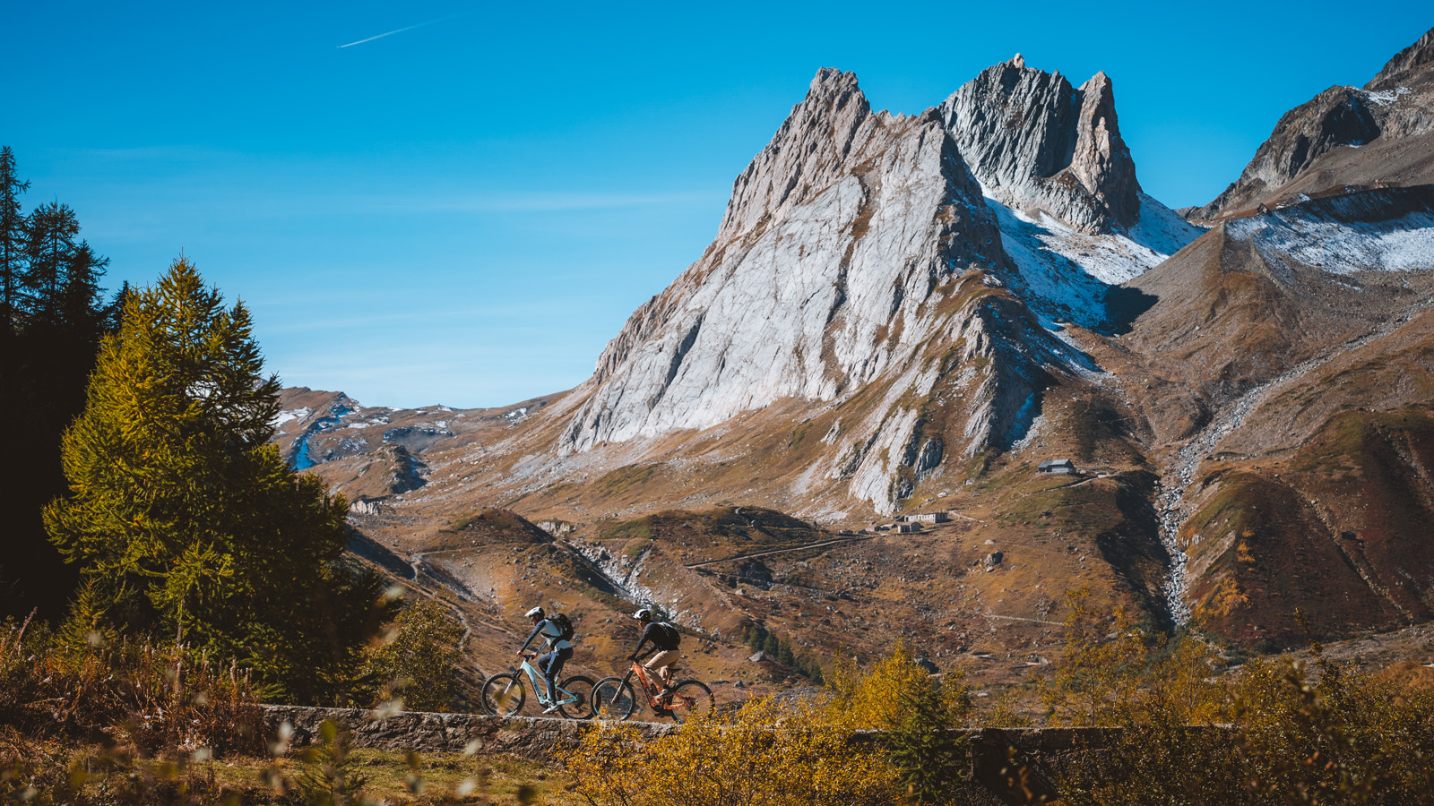 Two cyclists ride along a mountain trail under a clear blue sky. Majestic, rugged peaks rise in the background, and autumn trees frame the scene.
