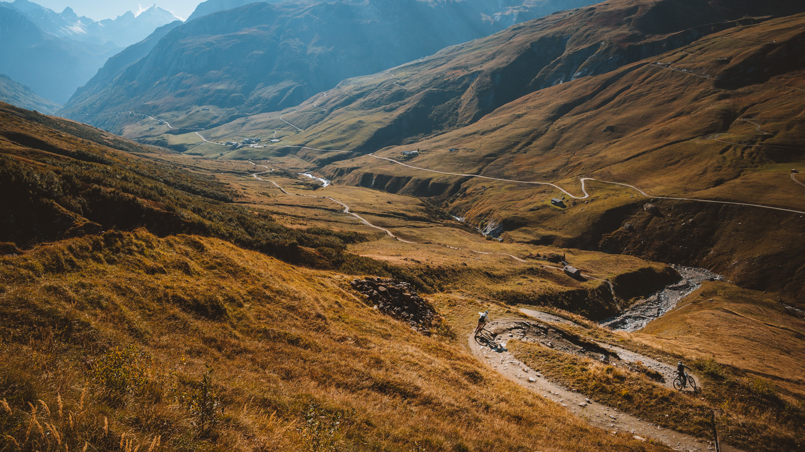 A biker navigates a winding trail through rolling green hills and mountains in a scenic landscape bathed in sunlight.