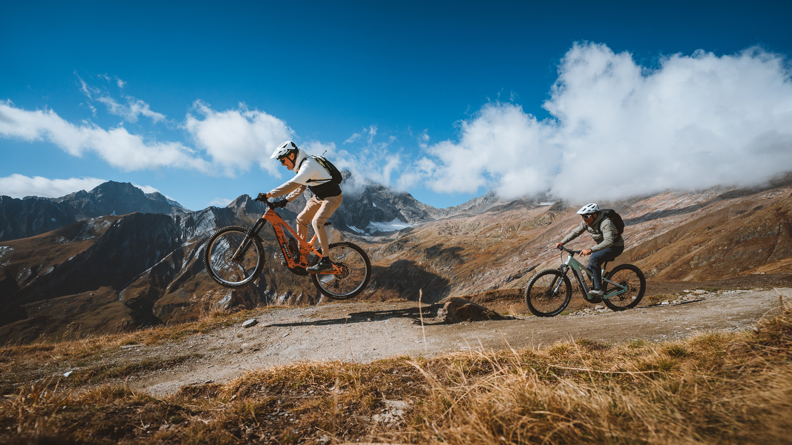 Two mountain bikers navigate a scenic trail in the mountains, with one airborne while the other rides steadily.