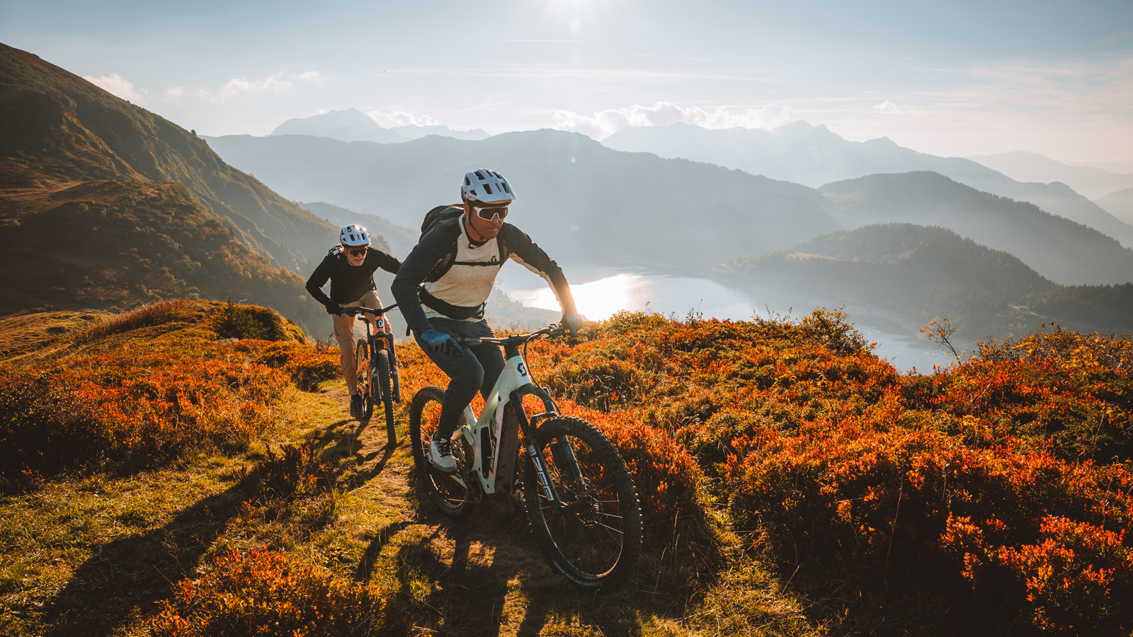 Two mountain bikers navigate a scenic trail through vibrant autumn foliage, with mountains and a lake in the background.
