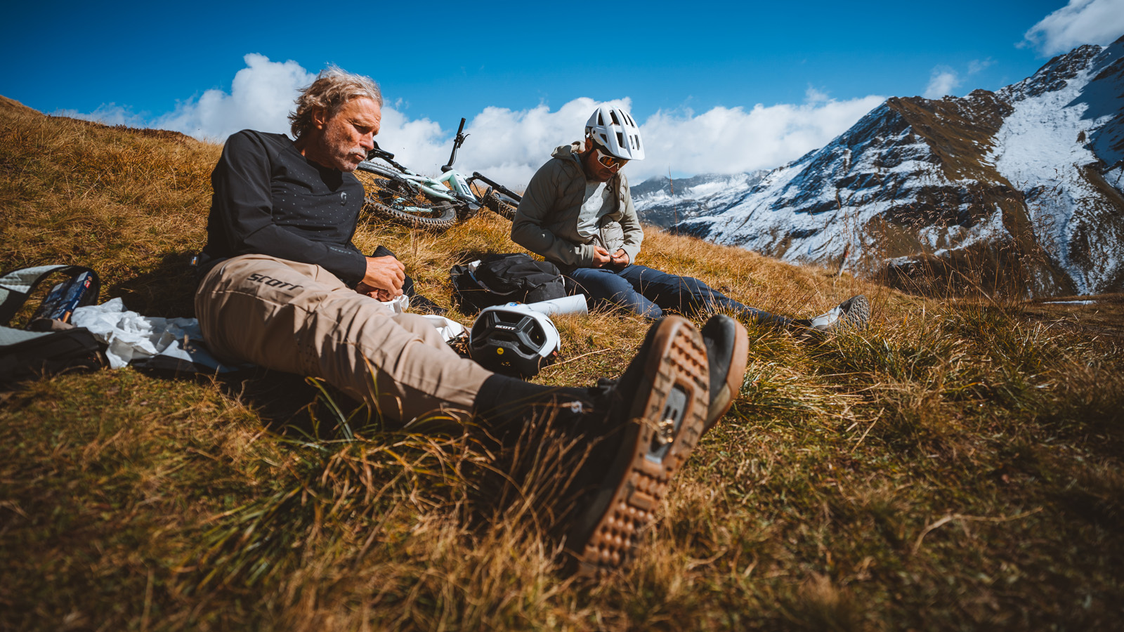 Two cyclists resting on grassy mountain terrain, with bicycles and snow-capped peaks in the background under a bright blue sky.