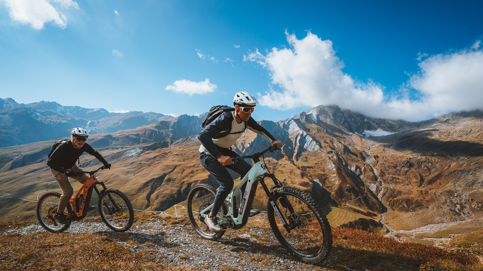 Two mountain bikers navigate a rocky trail against a backdrop of majestic mountains and blue skies, showcasing outdoor adventure.