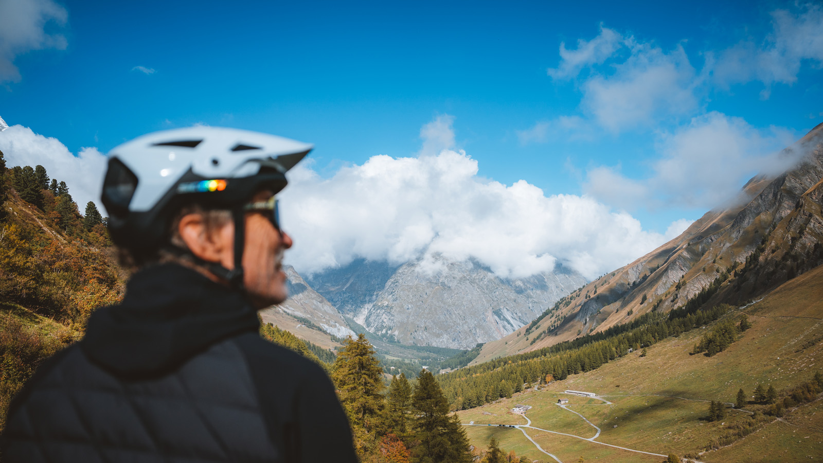 A cyclist wearing a helmet looks out over a sweeping mountain valley under a cloudy blue sky.