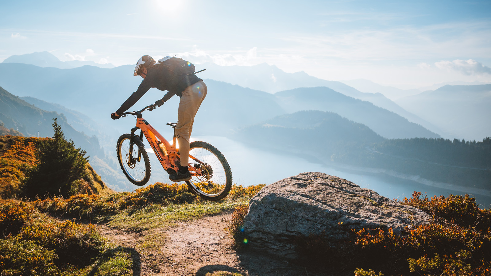 A mountain biker jumps an orange Scott Patron eRIDE mountain bike on a rugged trail overlooking a vast valley and lake.