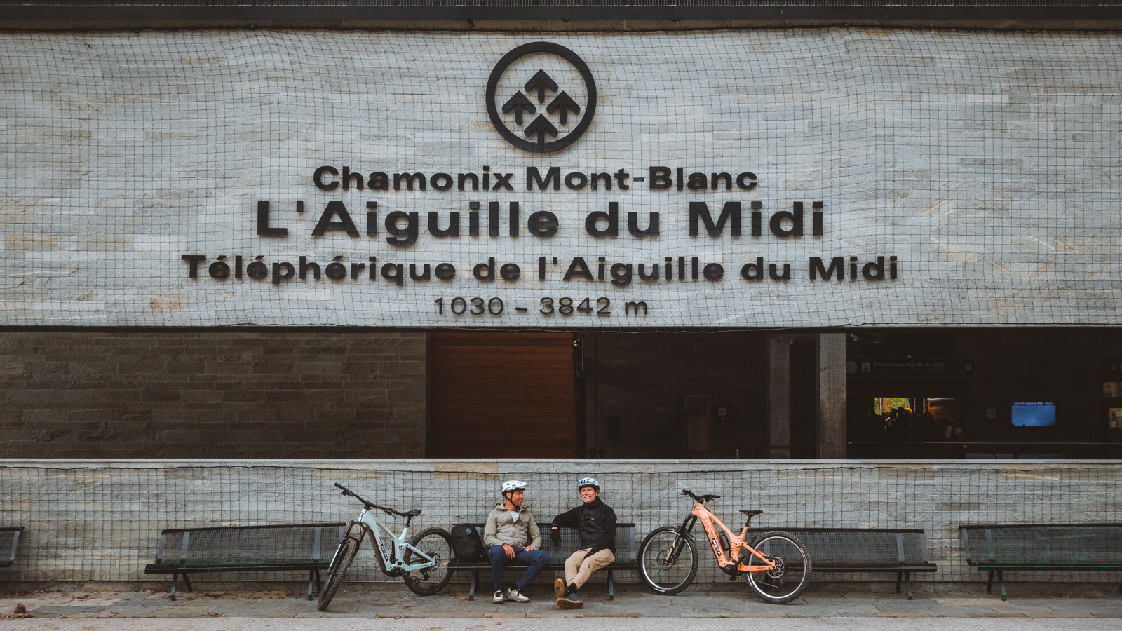 Two cyclists in helmets sit on a bench with their mountain bikes in front of the L’Aiguille du Midi cable car station in Chamonix.
