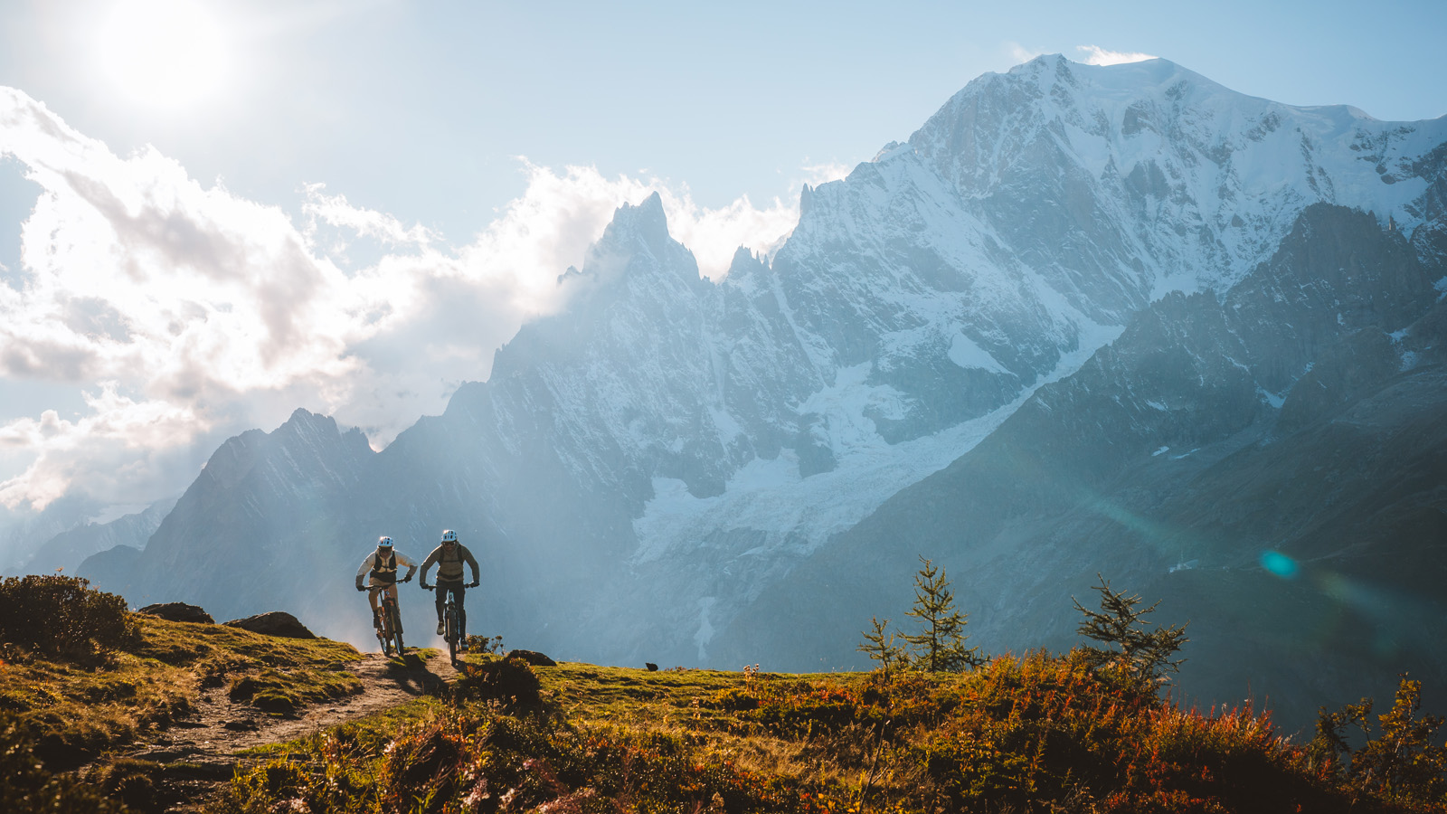 Two cyclists ride along a ridge toward the towering, snow-dusted peaks of the Mont Blanc massif under a bright, hazy sun.