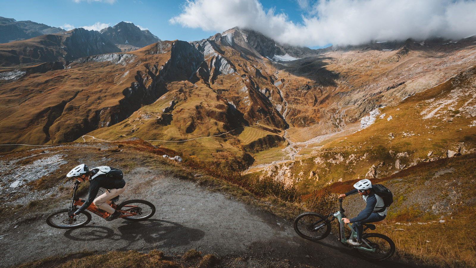 Two mountain bikers ride along a rugged trail overlooking a wide, golden alpine valley and jagged mountain peaks.