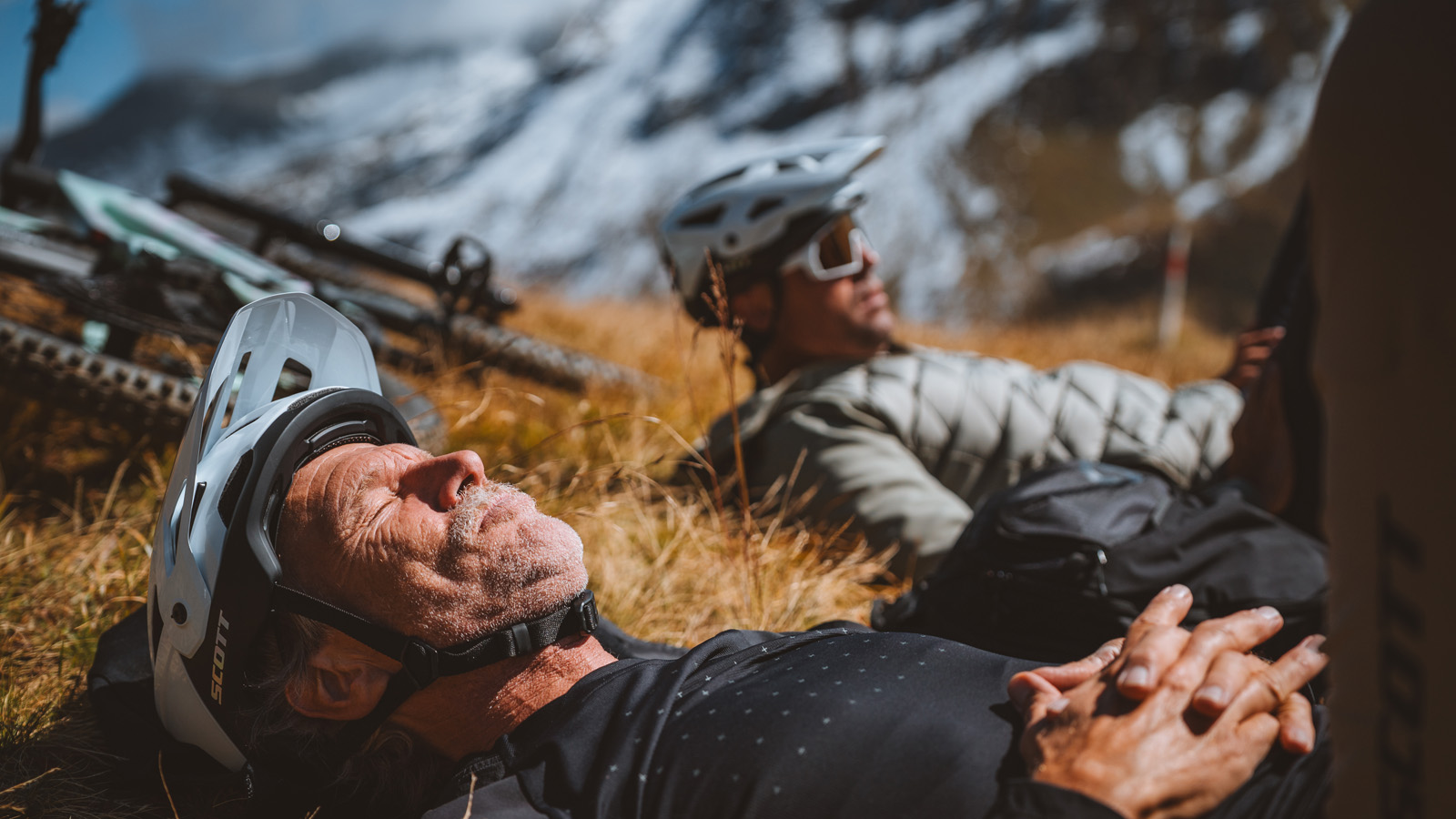 Two cyclists taking a break, lying in dry grass with their helmets on, with snow-capped mountains in the background.