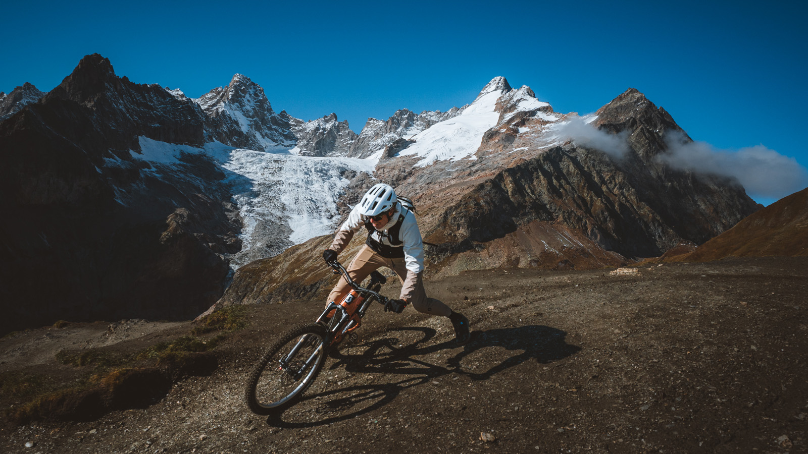 A mountain biker in a white helmet and jacket leans into a sharp turn on a rocky trail, set against a backdrop of massive, snow-covered peaks and a clear blue sky.