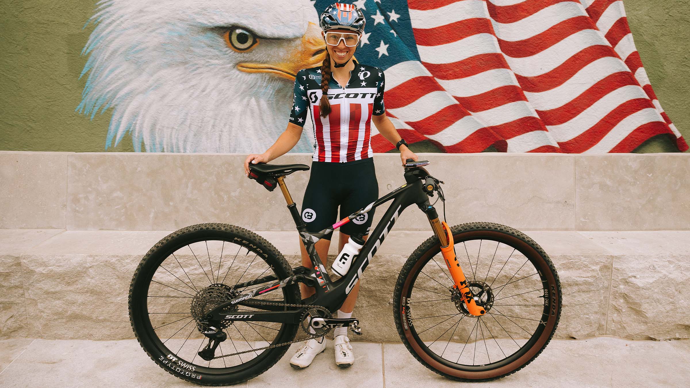 Hannah Otto stands smiling behind a SCOTT Spark RC mountain bike, wearing the XCM National jersey and USA themed helmet, posed in front of a mural featuring a bald eagle and the American flag.