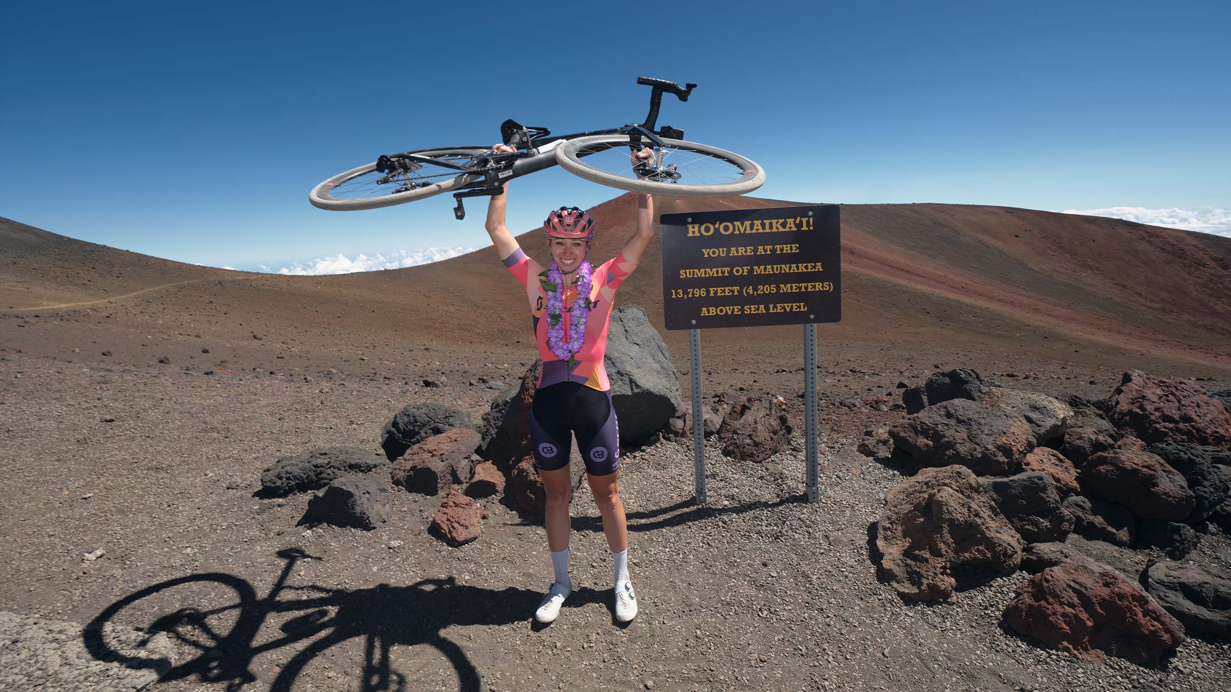 Hannah Otto celebrates at the summit of Mauna Kea, Hawaii, holding her bike overhead beside a sign marking 13,796 feet above sea level, surrounded by rocky volcanic terrain under a clear blue sky.
