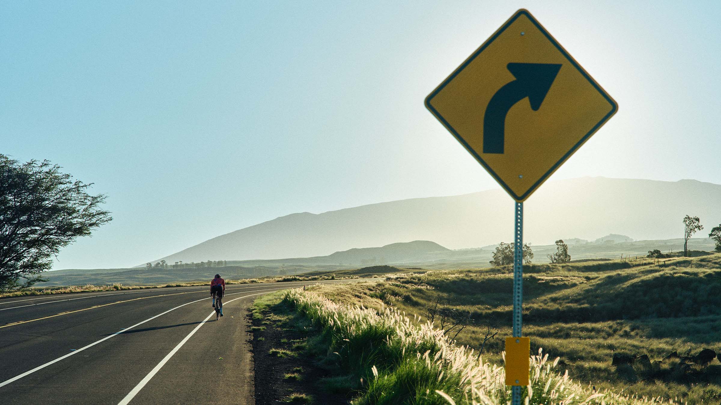 Hannah Otto rides solo along a quiet Hawaiian road into a challenging headwindduring her FKT attempt, passing a right-turn road sign with rolling hills and misty mountains in the background.