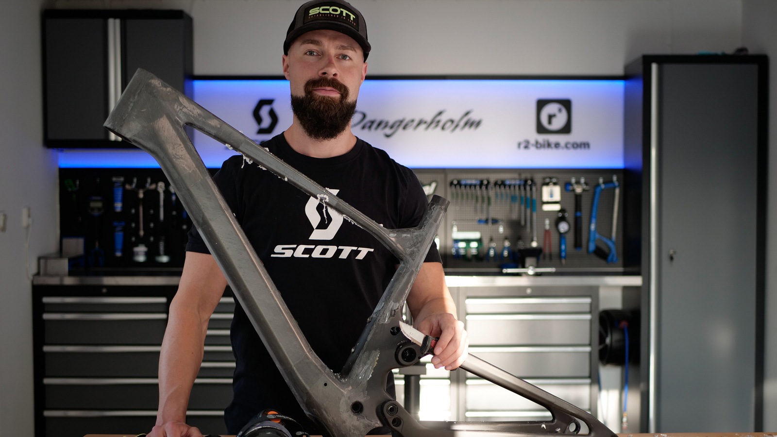 Gustav Gullholm standing next to a dark green Scott mountain bike with beige tires, leaning against a white and gray wall.