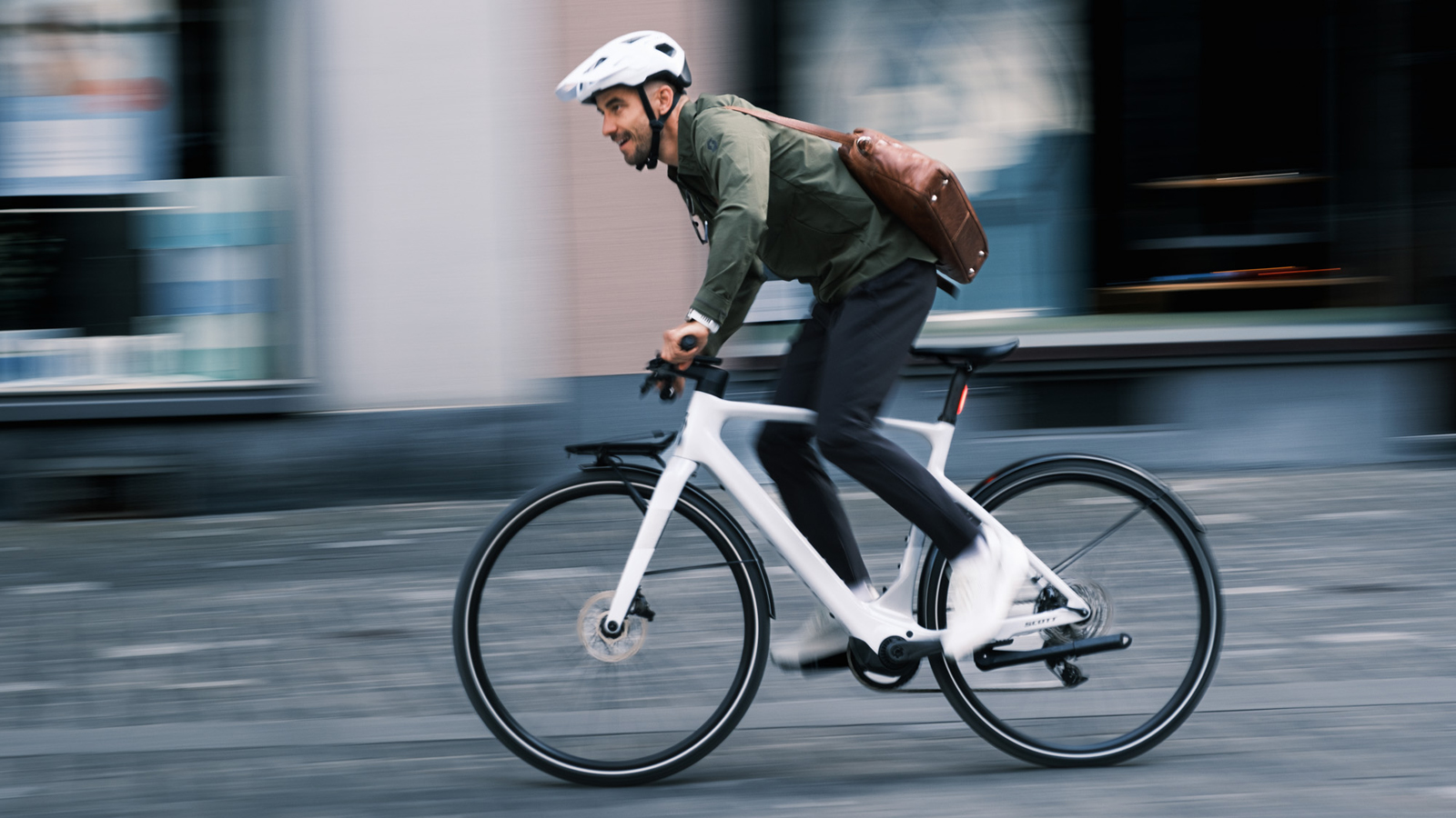 Nino Schurter is riding the SCOTT Silence eRIDE at speed in an urban environment. The SCOTT Silence eRIDE has an integrated frame, disc brakes, and thin tires designed for city use. Nino Schurter is dressed in a green shirt, black pants, white sneakers, and a brown leather shoulder bag. The background appears blurred due to motion, with storefronts visible in the distance.