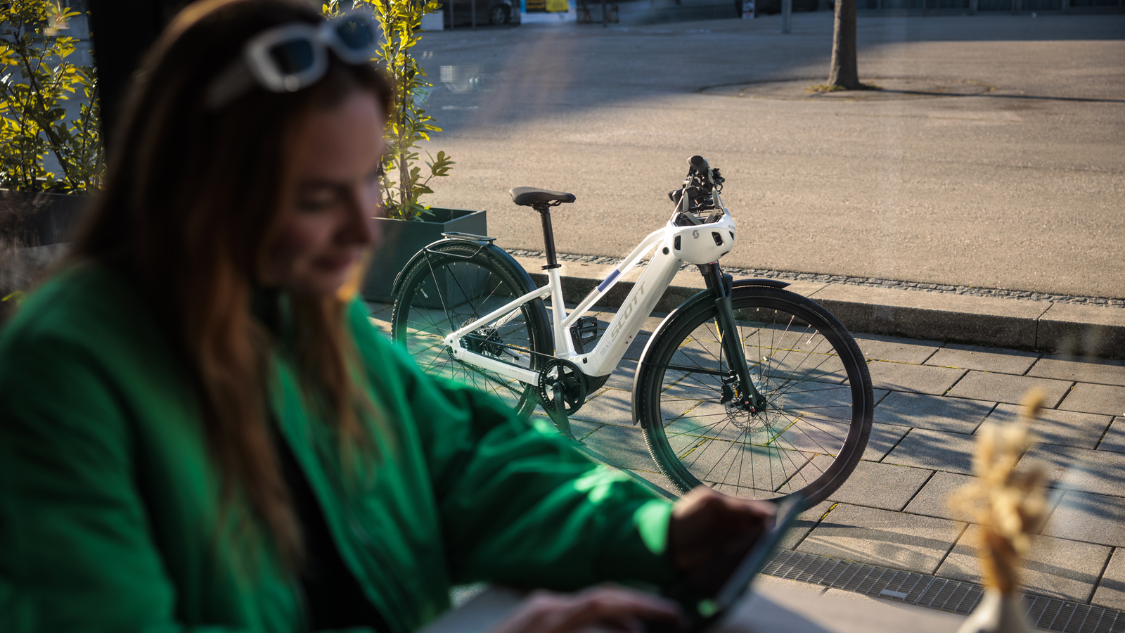 A white SCOTT Passage bike parked outdoors near a café, with a person seated inside wearing green clothing and sunglasses.