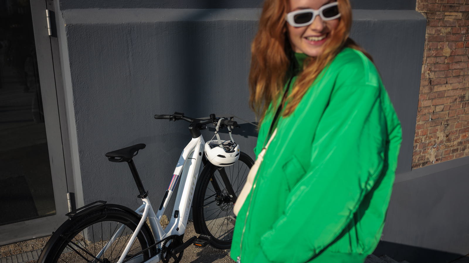 A smiling woman in a bright green jacket and white sunglasses stands in the foreground, slightly out of focus. Behind her, a white Passage SCOTT Bike with a black seat, handlebars, and a rear rack is parked against a gray wall. A white helmet hangs from the bike's handlebars. The background includes a glass door and part of a brick wall.
