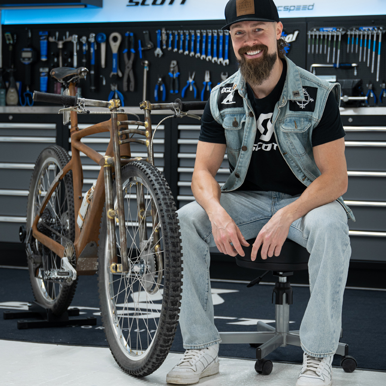 Gustav Gullholm seated in a workshop next to a custom wooden mountain bike with brass accents, surrounded by tools and SCOTT branding.
