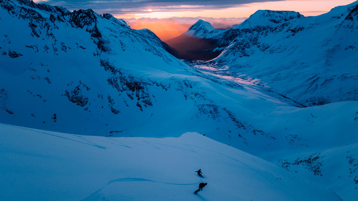 Sunset on mountains with two snowboarder riding white powder