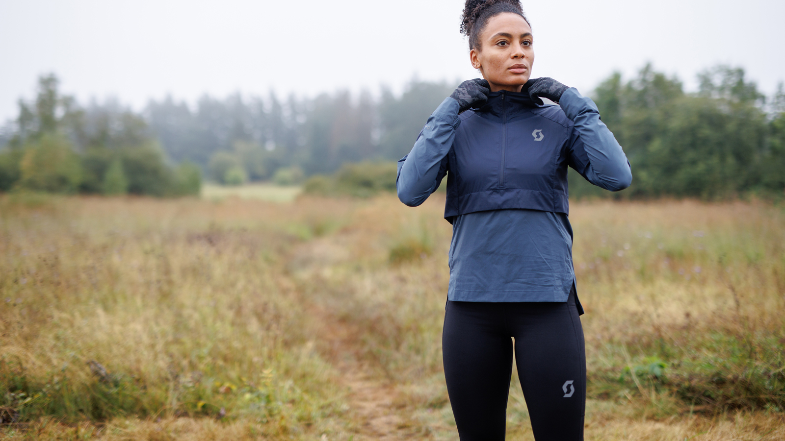 Woman in winter running gear adjusting her jacket on a scenic trail. Dressed in a navy blue windproof jacket, black leggings, and gloves, she prepares for a cold-weather outdoor run. Perfect for fitness, sportswear, and active lifestyle themes.